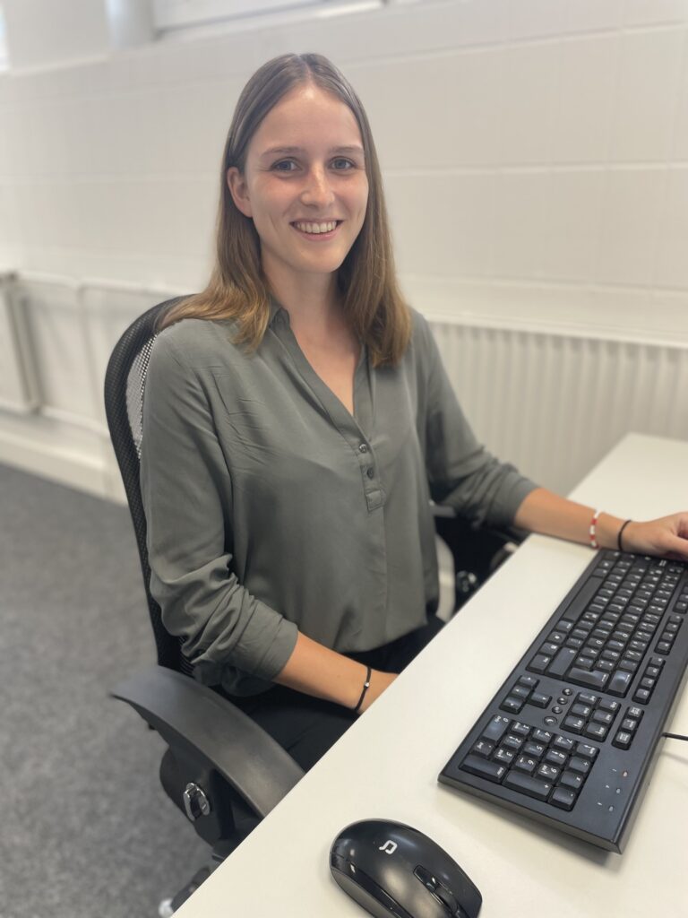 Lächelnde junge Frau mit braunen Haaren in grauer Bluse sitzt an einem weißen Schreibtisch mit Tastatur und Maus in einem hellen Büro.