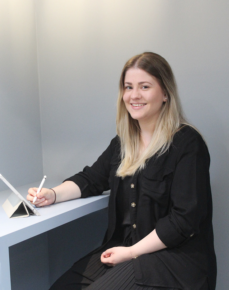 Lächelnde junge Frau mit langen blonden Haaren in schwarzer Kleidung sitzt an einem Tisch und hält einen Stift über ein aufgestelltes Tablet.